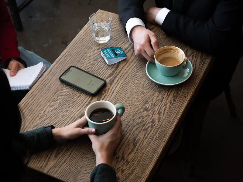 Two people sitting across a wooden table with cups of coffee, mid-conversation.